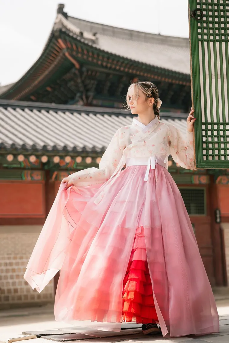 Woman in a pink and white hanbok holding out her skirt beside a palace hall