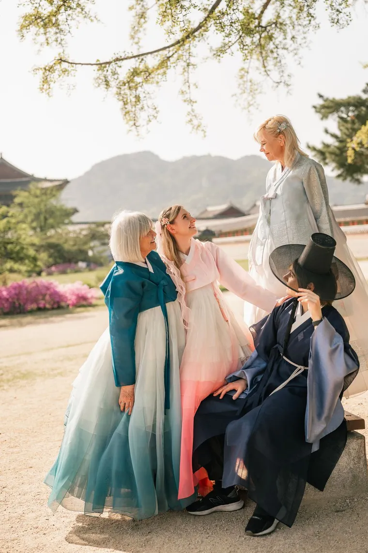 Three friends in pastel hanbok with a man in a gat, laughing on palace steps