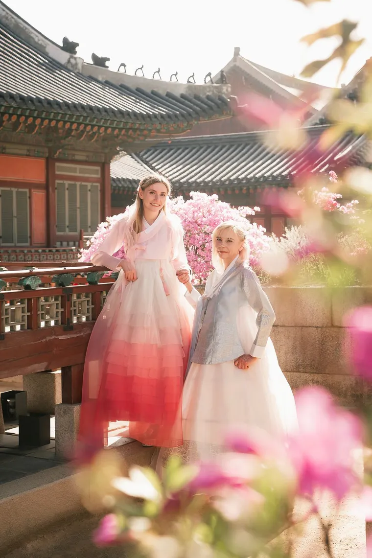 Two friends in pink and white hanbok standing at a palace railing among azaleas