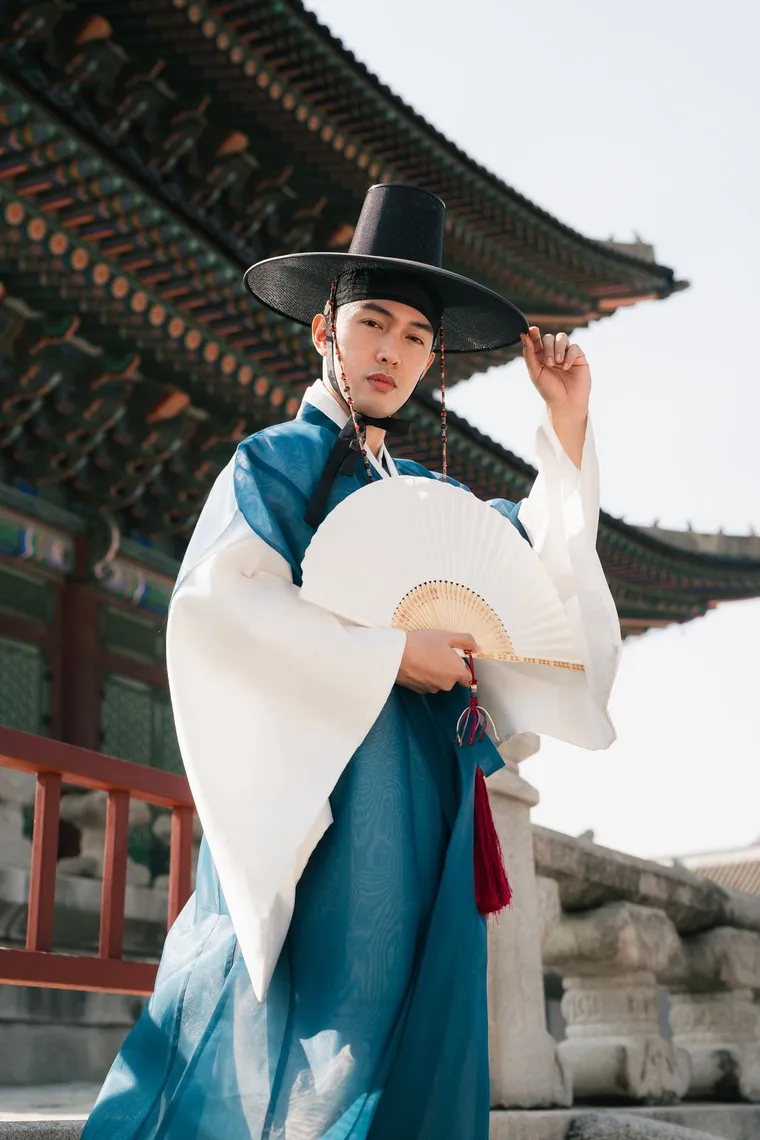 Man in a blue hanbok and gat holding a folding fan under a palace roof