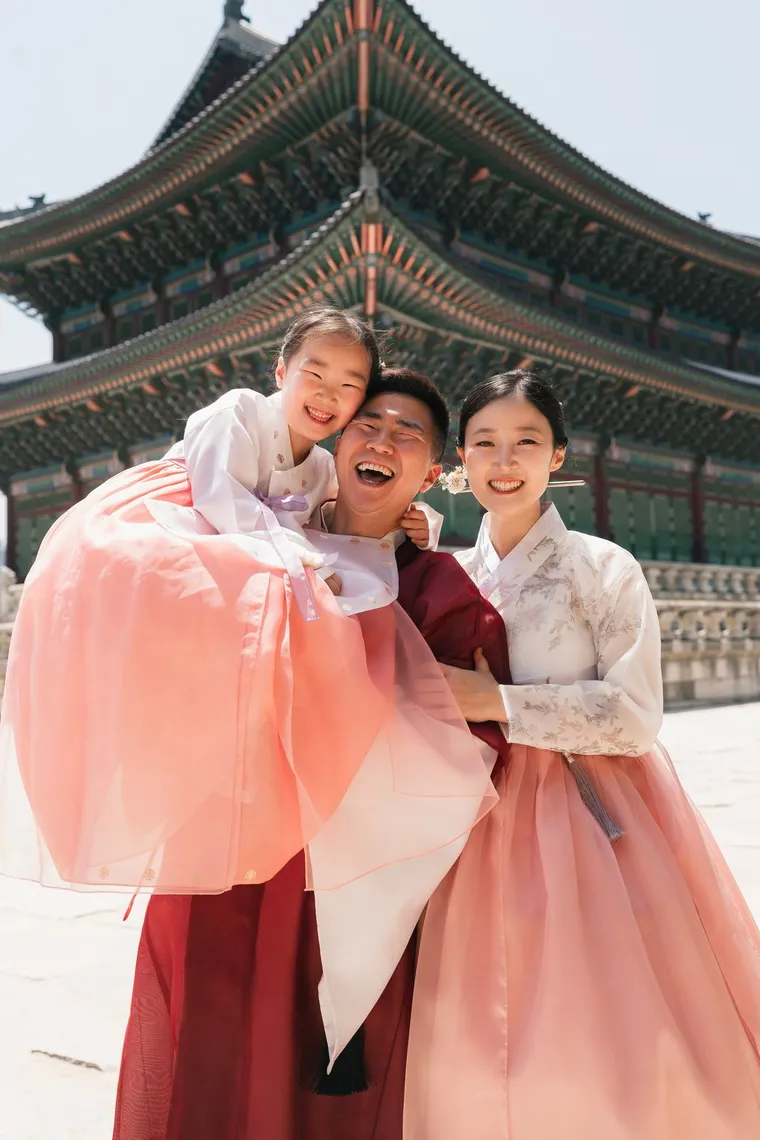 Family in pink and crimson hanbok laughing together in a palace courtyard