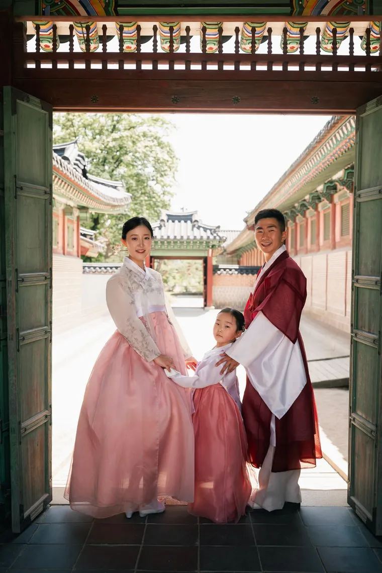 Family of three in hanbok walking hand in hand through a palace corridor