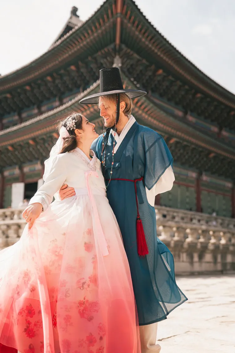 Couple in blue and pink hanbok embracing in a red-columned colonnade
