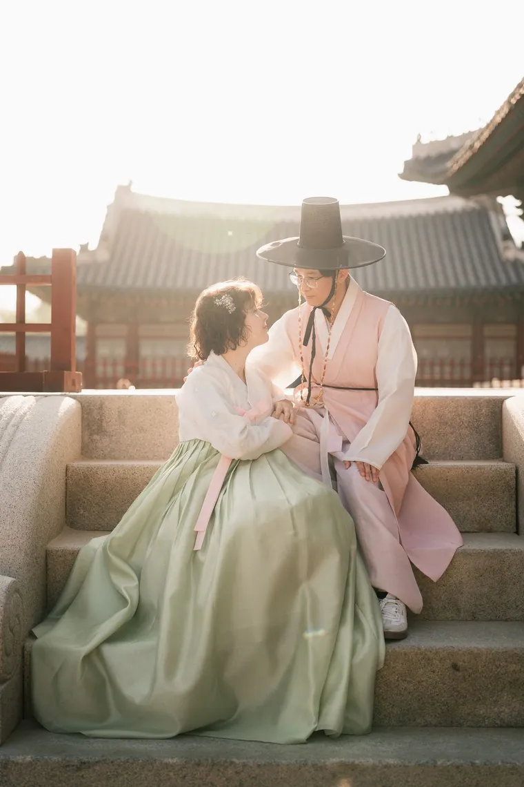 Couple in pastel hanbok sitting on palace steps, looking at each other