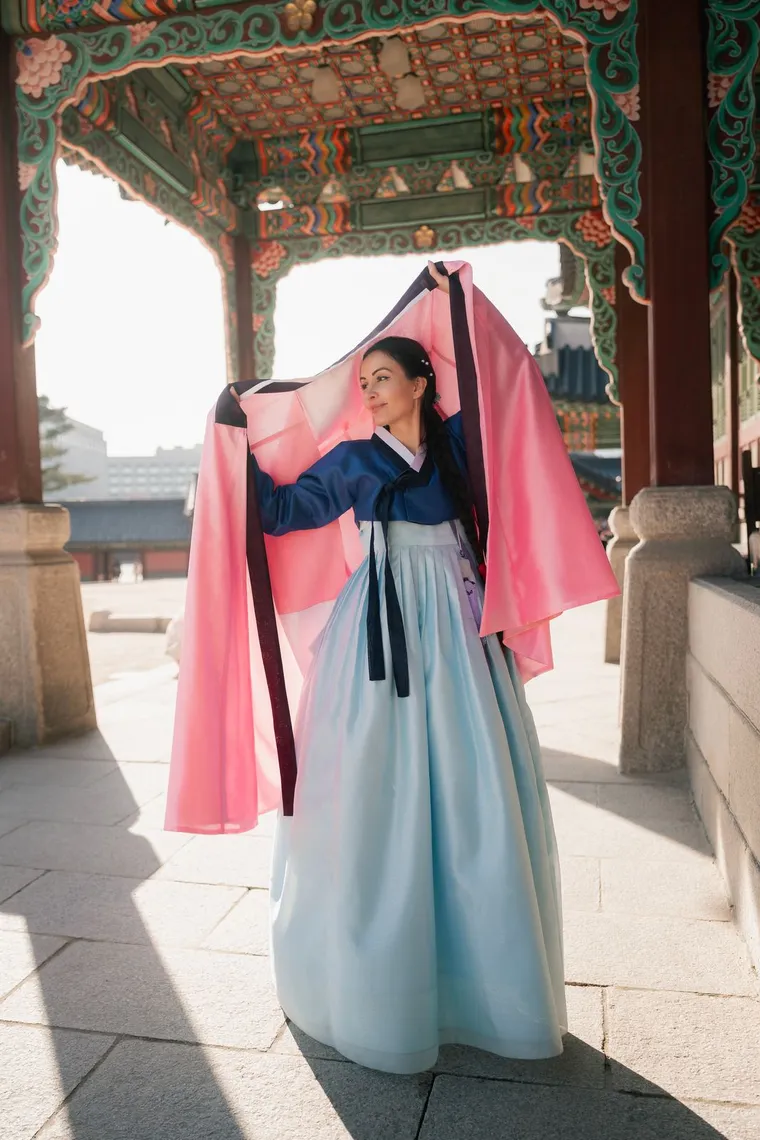 Woman in a light blue hanbok lifting a pink shawl overhead in a palace courtyard