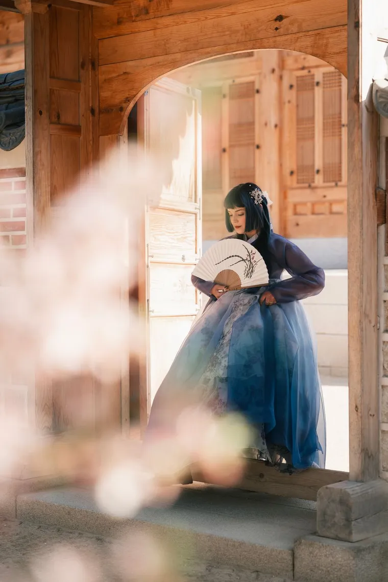 Woman in a blue hanbok holding a fan in a sunlit palace doorway