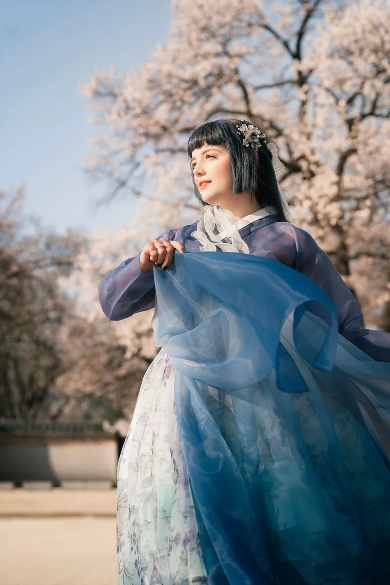 Woman in a blue hanbok lifting her skirt beside spring blossoms