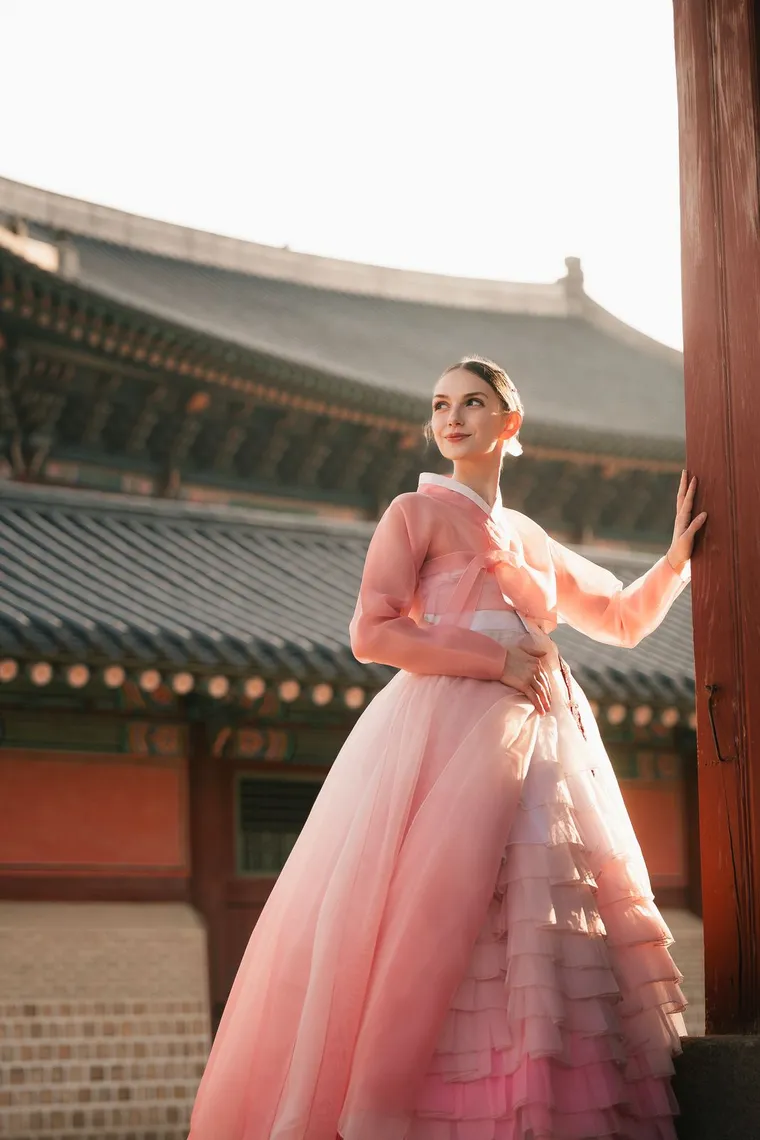 Woman in a pink hanbok on a palace terrace with roofs and mountains behind