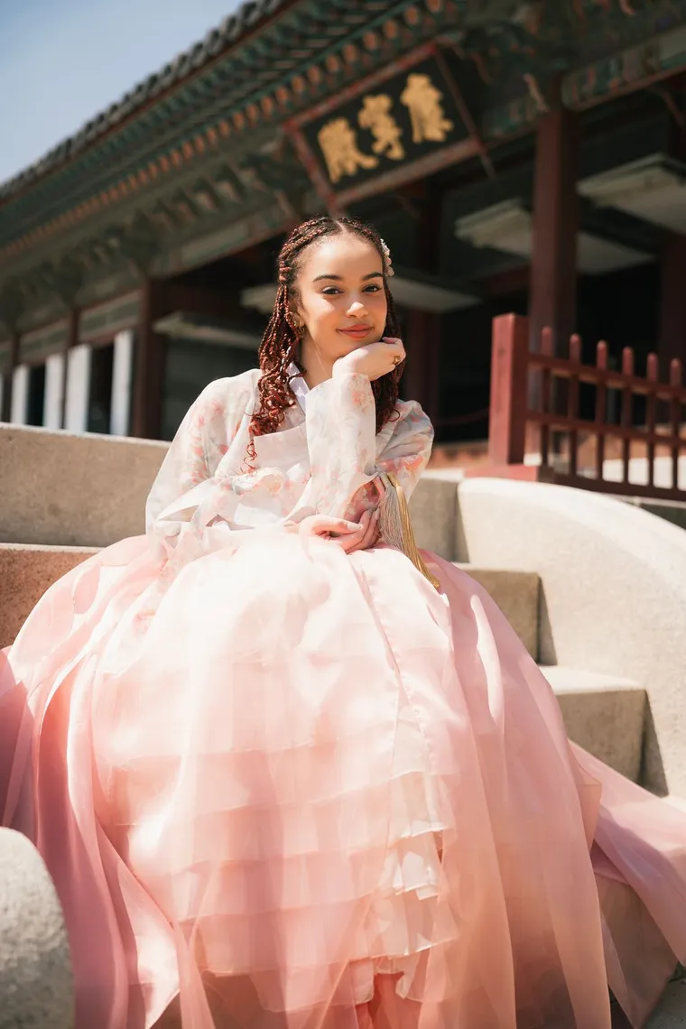 Woman in a white and pink hanbok seated on palace steps, chin on hand