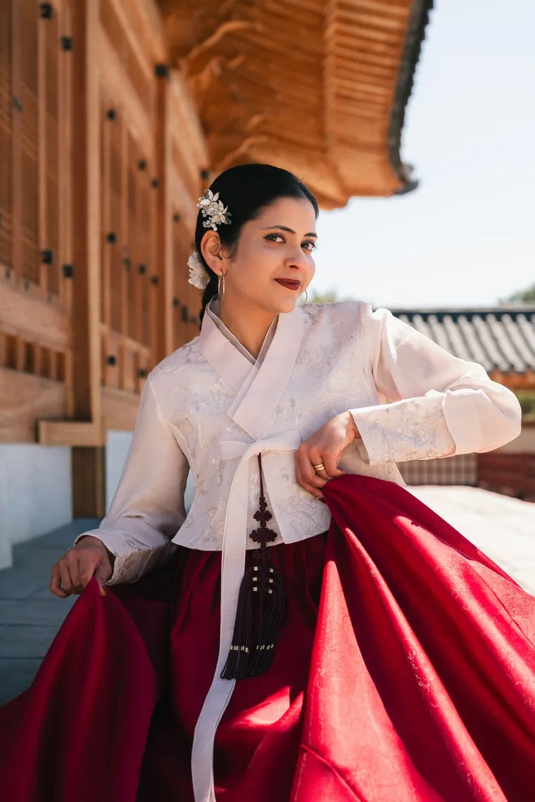 Woman in a white and crimson hanbok holding her skirt in a colonnade