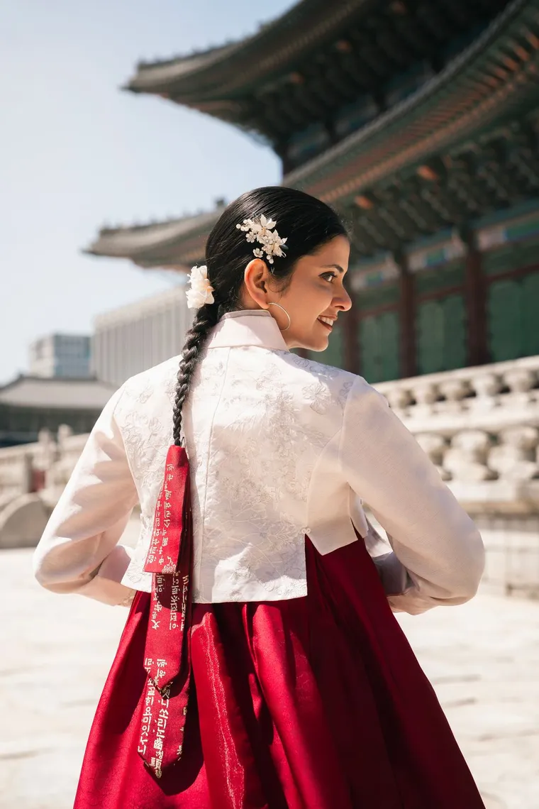 Woman in a white and crimson hanbok seen from behind, hair braided with flowers