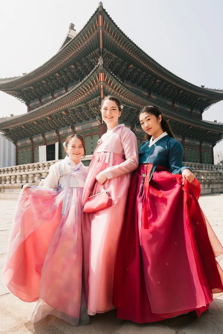 Three friends in pink and navy hanbok holding out their skirts in front of a palace hall