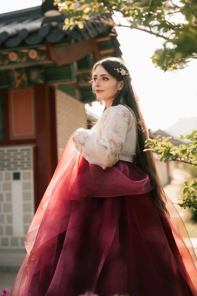 Woman in a white and crimson hanbok holding her skirt beside a palace wall