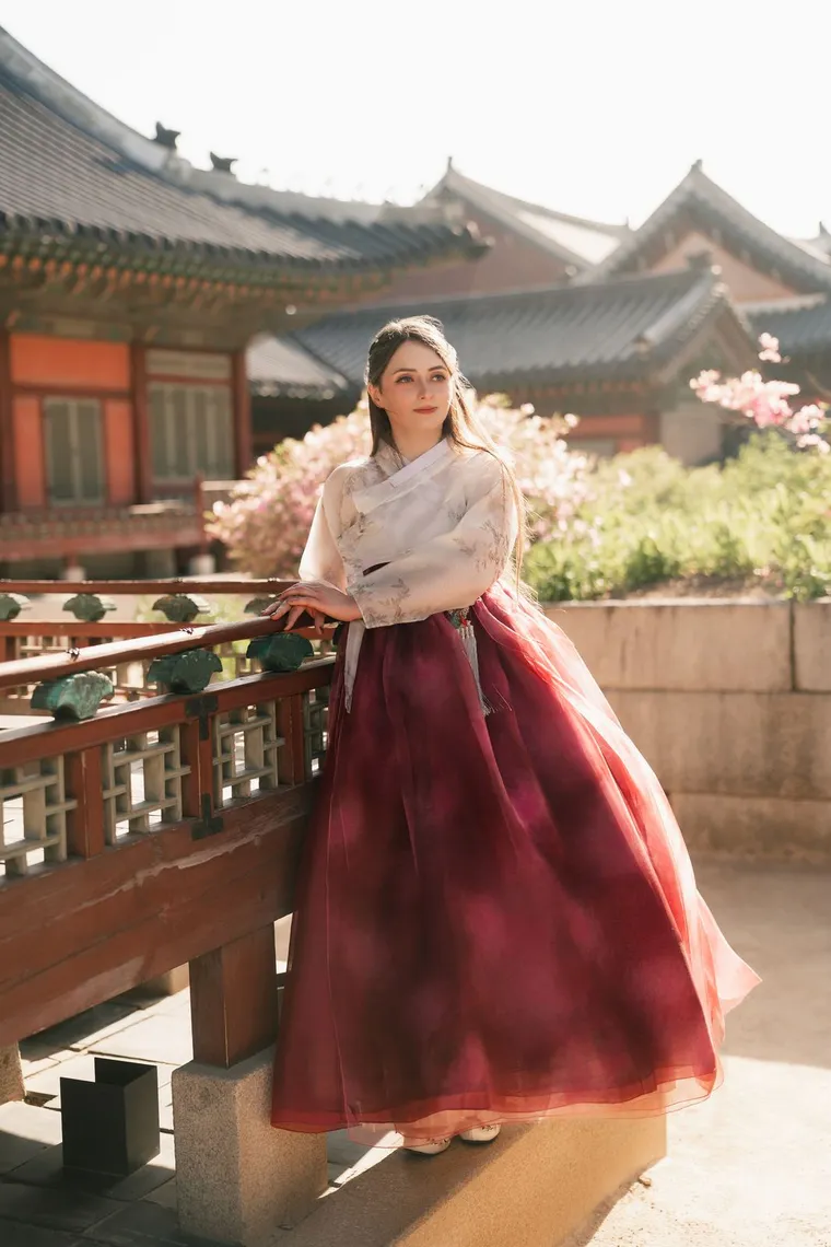 Woman in a white and crimson hanbok at a palace railing among blossoms