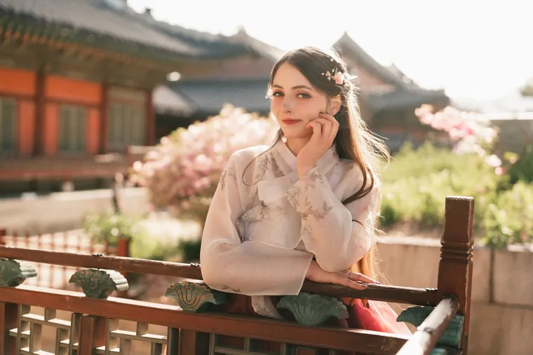 Woman in a white hanbok smiling at a palace railing, blossoms behind