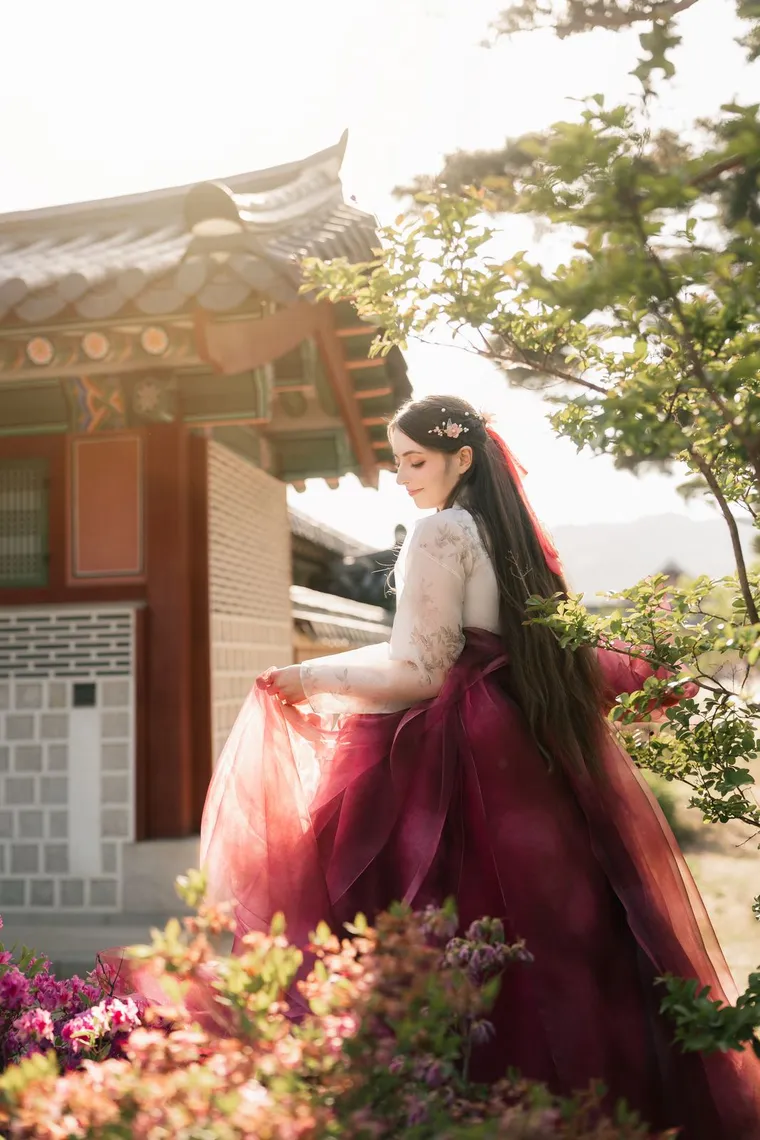 Woman in a crimson hanbok skirt turning beside a hanok wall