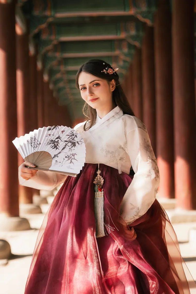 Woman in a white and crimson hanbok holding a painted fan in a colonnade