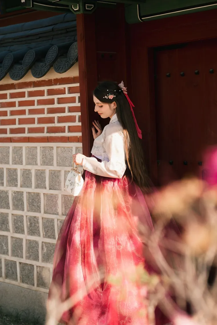 Woman in a crimson hanbok leaning against a patterned palace wall