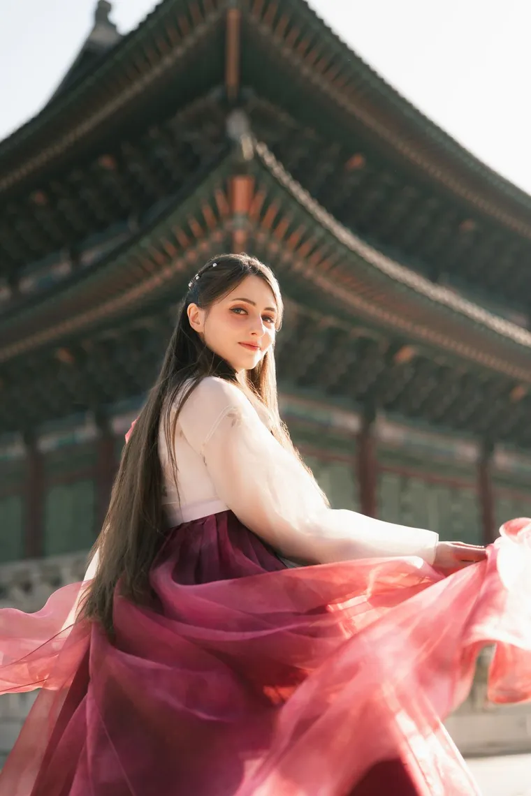 Woman in a white and crimson hanbok seated in a palace corridor