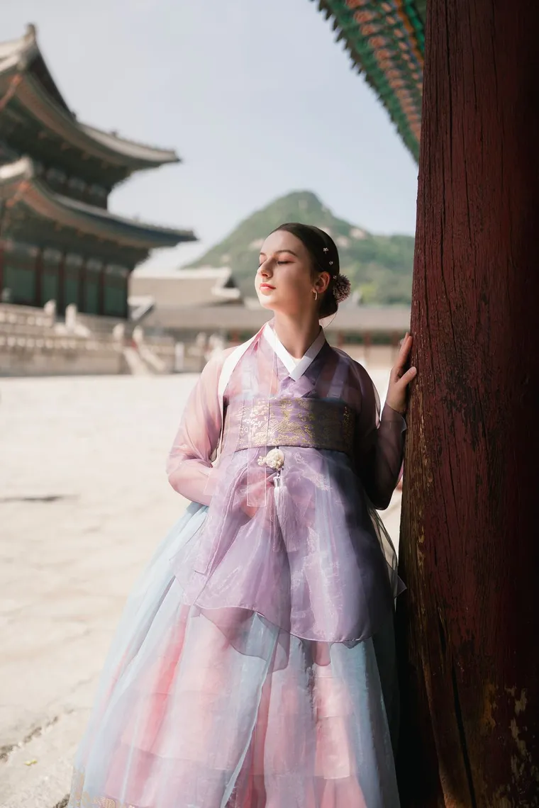 Woman in a lilac hanbok beside a palace column, mountain behind