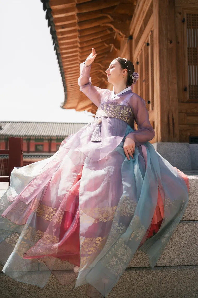 Woman in a lilac hanbok seated on a palace stair with her arm raised