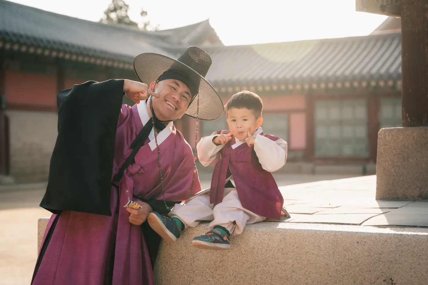 Father and small son in hanbok sitting together on palace steps, both laughing